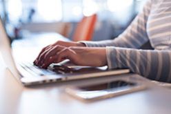 一名身穿条纹衬衫的女子在笔记本电脑上打字，手机就放在旁边。